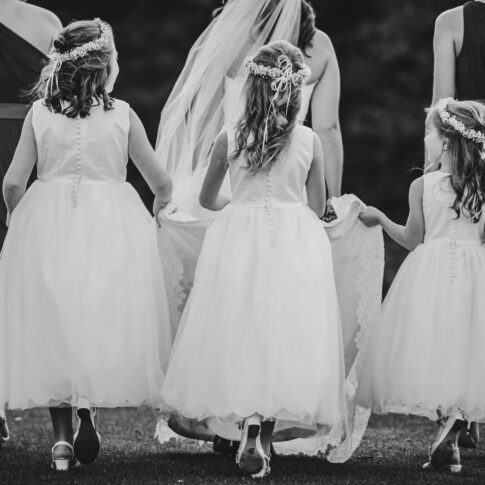 Three flower girls all assist in lifting the brides hem as she walks across grass with her bridesmaids.