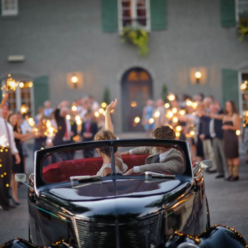 A bride and groom wave goodbye to their guests who are waving sparklers as they drive away from their reception at Hotel Domestique.
