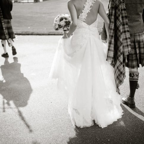 A bride holds her groom's hand as they follow a bagpiper towards their reception venue with their wedding party following closely behind them.